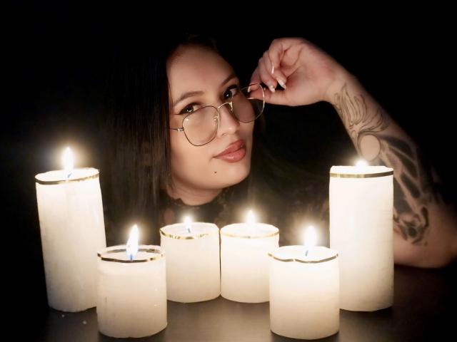 LiliethWolf with glasses, dark hair, and tattoos, lying by glowing white candles, gently touching her nose. Her ass is partially visible under her.