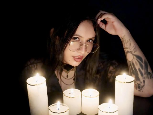 LiliethWolf with glasses, tattoos, and a choker, holding her hair, surrounded by lit candles, has a sexy, moody expression, and a small beauty mark on her cheek.