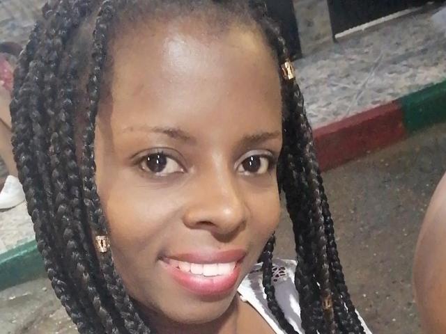 CheatingNeighbor has dark brown skin, medium-length black braids, brown eyes, pink lipstick, and a small smile. She's wearing a white shirt with a black pattern.