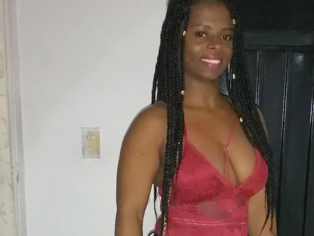 CheatingNeighbor with dark skin, long braided hair, wearing a red lace dress, showing her big boobs and smiling. She's standing in a room with a black door.