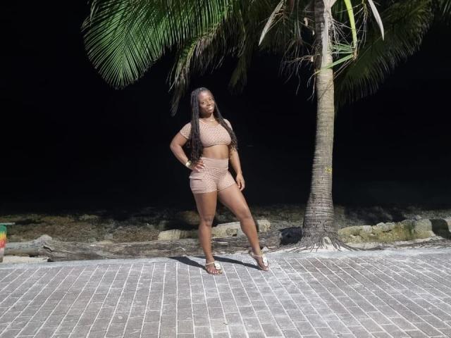 CheatingNeighbor with dark skin, long black hair, big ass, and curvy figure, wearing a beige crochet bikini, standing on a brick path, under a palm tree at night.