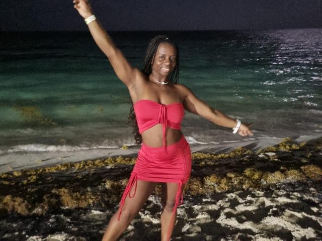 CheatingNeighbor with dark skin, long braids, and a red strapless top and skirt, raising one arm, smiling, standing on rocky beach at night, wearing a white bracelet.