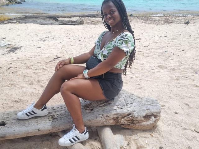 CheatingNeighbor with dark skin, long braids, green leaf pattern top, black shorts, white Adidas sneakers, sitting on a log, smiling, beach background, tan legs, black ass.