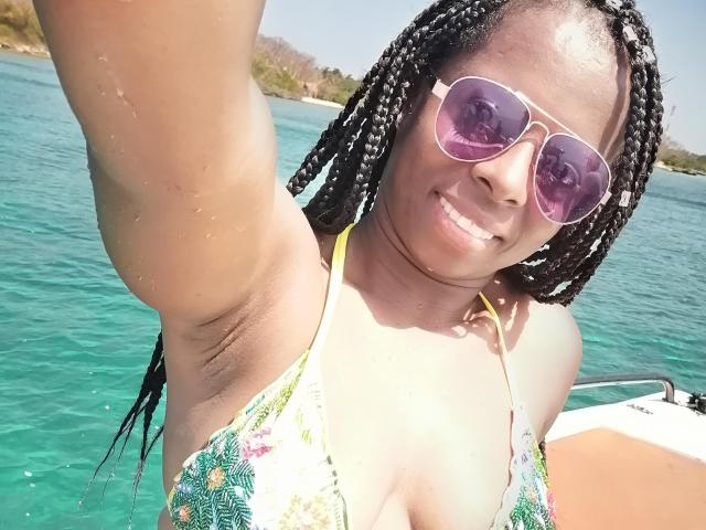 CheatingNeighbor with dark skin, braided hair, wearing pink sunglasses and a floral bikini, showing tits in a selfie on a boat, smiling at the camera.