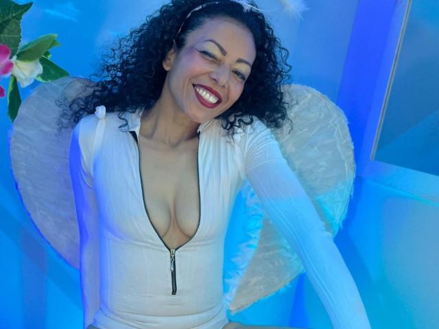 BrendaLive with curly black hair, smiling, wearing a white zip-up dress with deep cleavage, exposing her tits, blue light background, fluffy white wings, and red lipstick.