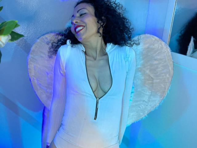 BrendaLive with curly black hair, smiling, wearing a white zip-up bodysuit with a deep cleavage, showing her tits, against a blue-lit wall with a fuzzy heart.