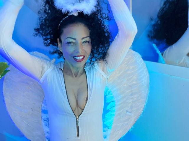 BrendaLive with curly black hair, white sheer top, deep cleavage, smiling, arms raised, wearing fluffy white headpiece, blue light, white wings, reflection in mirror.