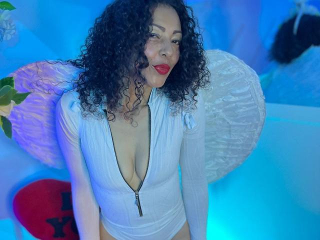 BrendaLive with curly black hair, red lipstick, white zip-up bodysuit showing her tits, blue lighting, paper lantern, and red heart-shaped pillow in the background.