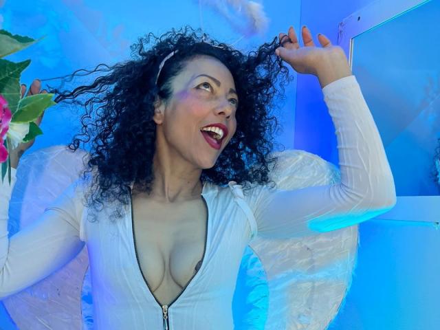 BrendaLive with curly black hair, white zip-up top, revealing cleavage, smiling, holding hair, blue lighting, white wings, pink flowers in left. Boobs, tits, top, smiling.