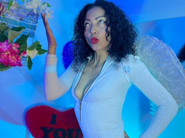 BrendaLive with curly black hair, red lipstick, white zip-up top, showing big tits, holding flowers, wearing angel wings, against blue light, hand on hip, looking up.