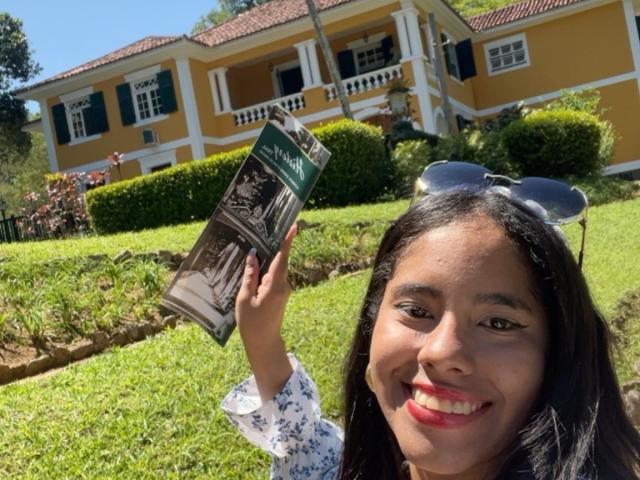 SiennaTaylor with dark hair, red lipstick, and white floral shirt, holding a book, smiling, blue sunglasses on head, standing on green grass, yellow house with white trim in background.