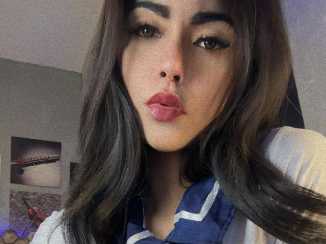 DangerousTrans69-hot with pouty pink lips, dark eyebrows, and long brown hair, wearing a blue and white top, looking directly at the camera. Boobs are not visible.