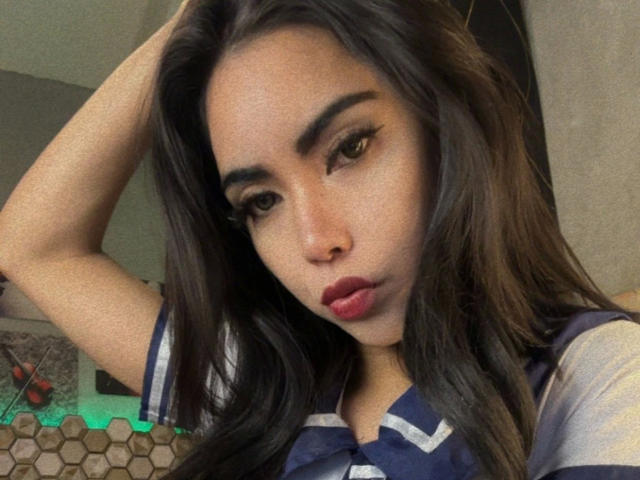 DangerousTrans69-hot with long dark hair, brown eyes, and red lipstick, wearing a blue and white striped shirt, looking down with a pouty expression, hand in hair.