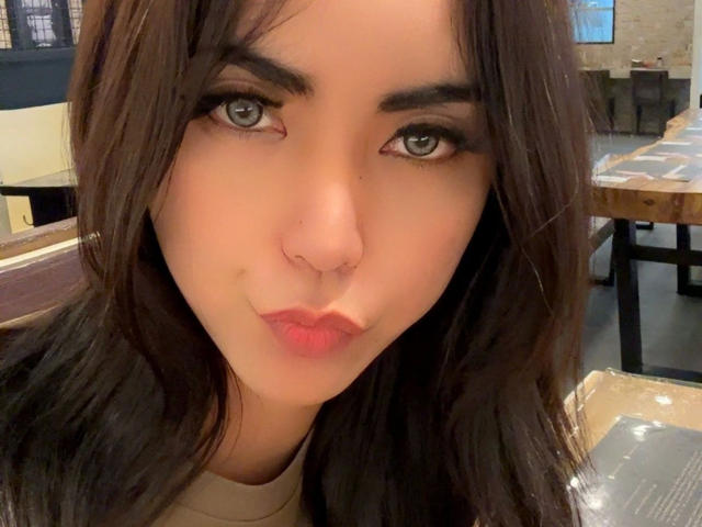 DangerousTrans69-hot with pouty pink lips, brown eyes, and long brown hair, giving a slight smirk. Boobless, no visible genitals. Soft lighting highlights her cute, feminine face.