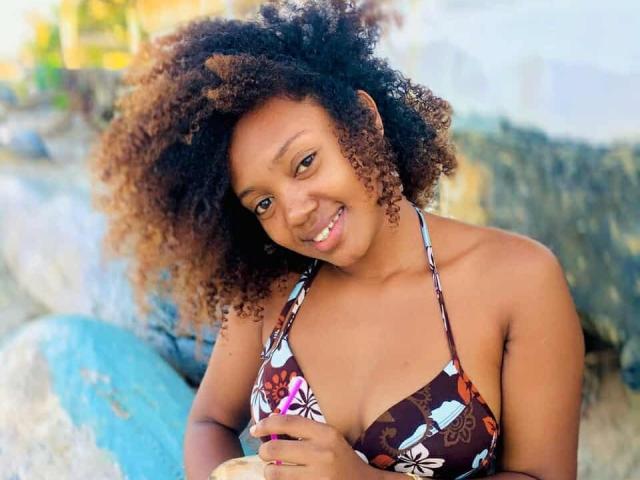 BarbaraSweetz with curly brown hair, smiling, wearing a brown floral bikini top, showing her tits, holding a blue drink, standing by rocks, beach background.