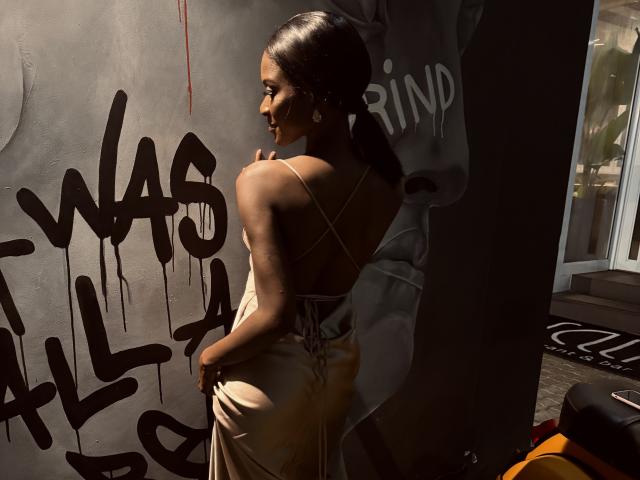 JodieFrancy with dark skin, long brown ponytail, wearing a backless, tan dress, standing against a graffiti-covered wall, sunlight highlighting her back and ass.