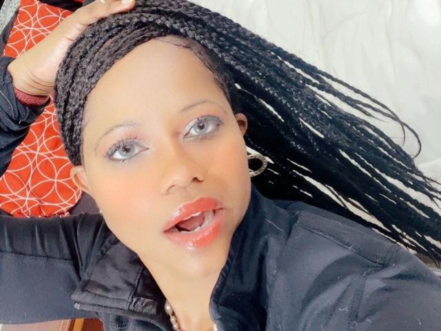 Cincia69 with long black braids, black jacket, shiny red lips, and hoop earrings, looking up with slightly parted red lips. Hand in braids, light skin, blue eyes, glistening face.
