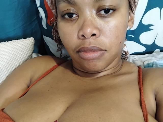WendyStacey with brown skin, medium-sized tits in an orange top, braids, and a serious expression, lying down. Her pussy is not visible.
