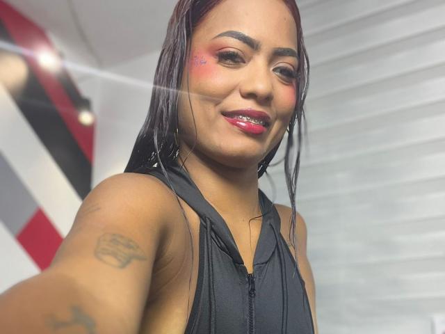 ChloeBrownOne with red lipstick, pink cheek blush, black halter top, long black hair, tattoo on right arm, smiling at camera, visible teeth, black eyeliner, white background.