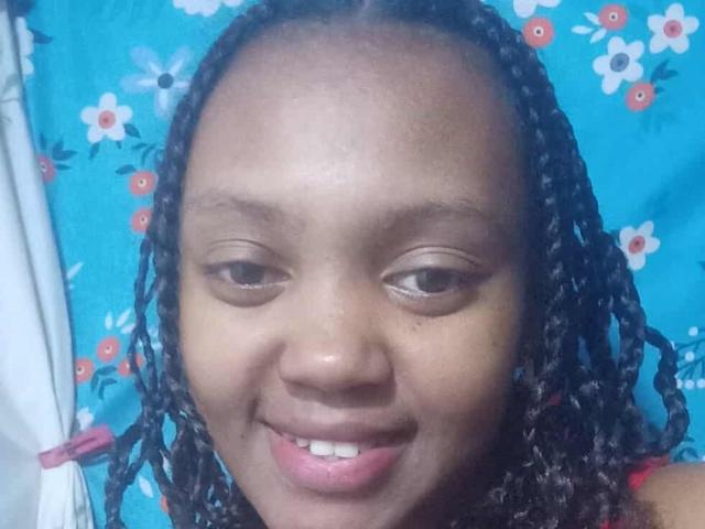 BarbaraSweetz with dark brown skin, small nose, and smiling, has braided black hair, and brown eyes, wearing a red top, against a blue floral background.