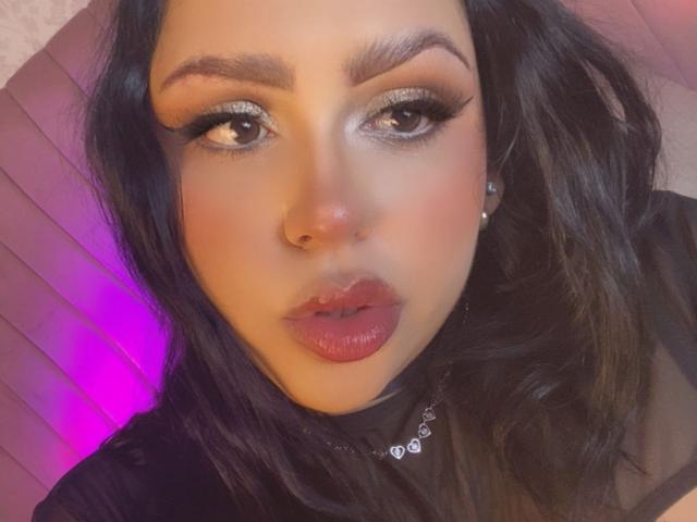 EmmaMyles with glossy pink lips, thick black eyebrows, shimmering eye makeup, and dark wavy hair, wearing a black top and diamond earrings, looking directly at the camera.
