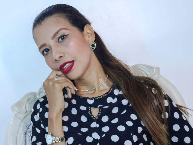 AnastasiaBlair with red lipstick, polka dot black top, pearl necklace, earrings, and ring, touching chin, smiling, long brown hair, white chair, light blue background, confident.
