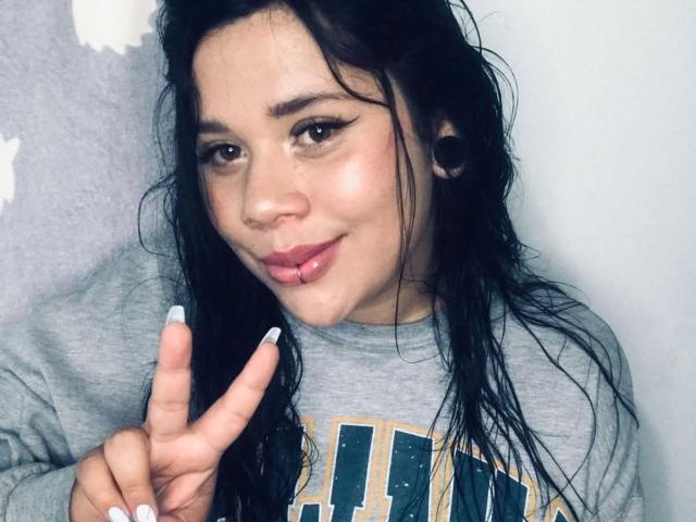 SamantaGreen with wet black hair, grey shirt, and nose ring, showing a peace sign with white nails, has a small pierced lip, and large black ear gauges.