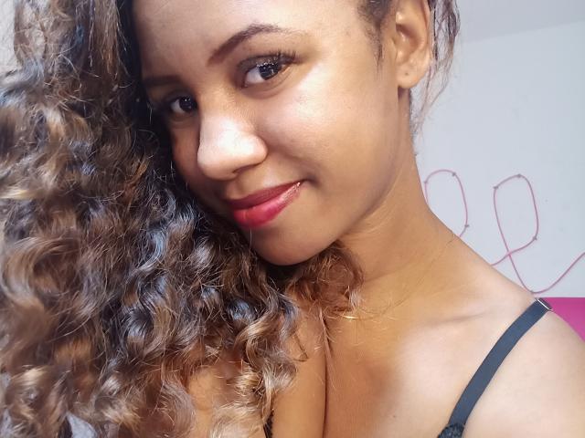 LayahCoquine has curly brown hair, light brown skin, and a cute smile with red lips. She's wearing a black strap, looking at the camera with a gentle expression.