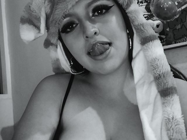 Black-and-white photo of Miyag with a fluffy striped headpiece, tongue out, heavy eyeliner, and a strapless top, showing her tits and a nose ring, looking seductive.