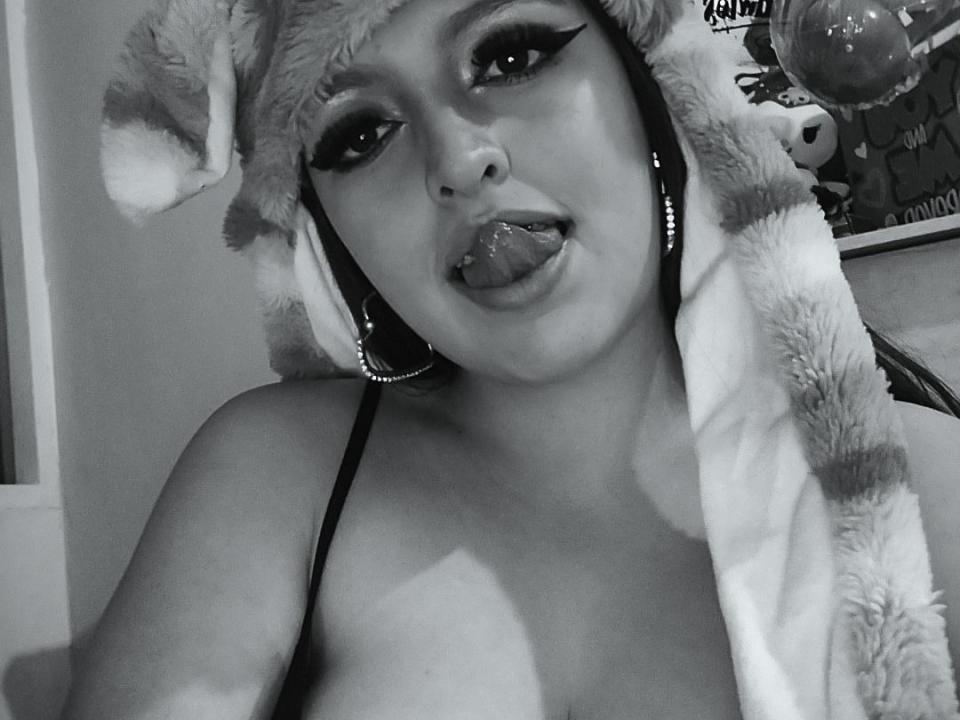 Black-and-white photo of Miyag with a fluffy striped headpiece, tongue out, heavy eyeliner, and a strapless top, showing her tits and a nose ring, looking seductive.