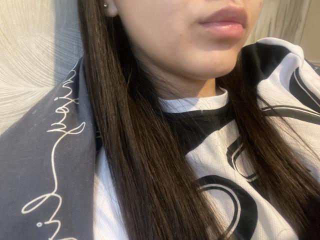 Beautifully69's face, showing glossy pink lips, straight brown hair, and a white shirt with black swirls. Her ear has a small stud earring.