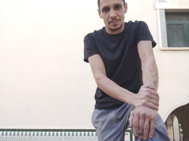 CabronAzos, wearing a black t-shirt and gray pants, sits with arms crossed, showing a tattoo on his right forearm, and a slight mustache. He looks relaxed.