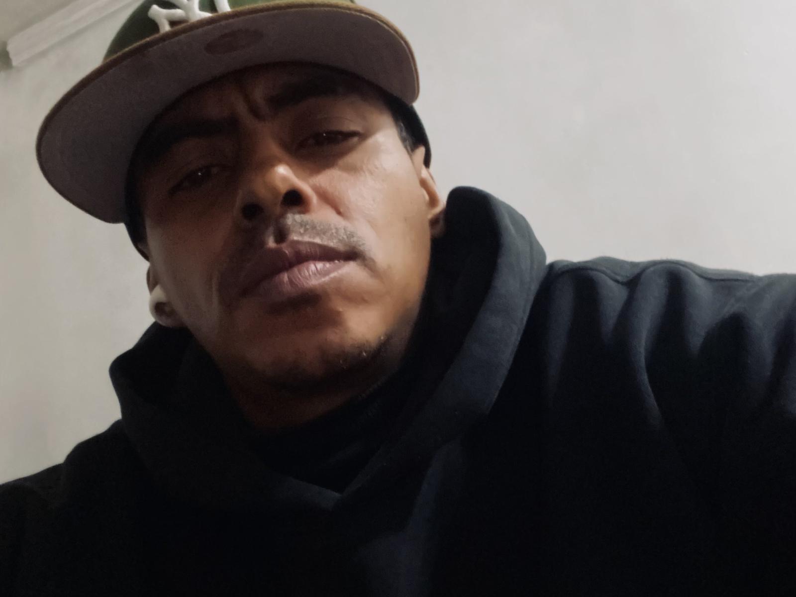 Indiopauzudo69, a medium-skinned man with a slight mustache, wearing a green hat and black hoodie, looking down with a serious expression. His nose is slightly upturned.