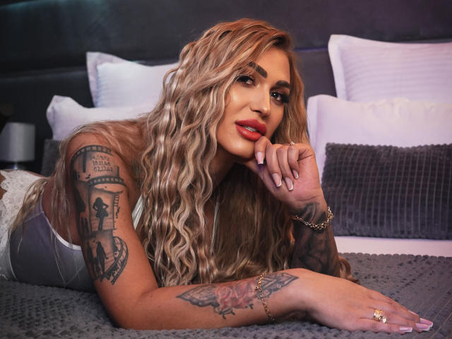 NicolSNaya met lang, golvend blond haar, rode lippenstift, tatoeages op de armen, liggend op een bed, een grijze, gestructureerde deken, grijze broek, een gouden armband, een ring, hand op het kin, sexy, zelfverzekerd..