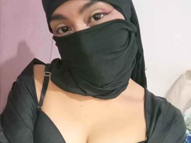 LaylaNoor with a black headscarf and mask, showing brown eyes with pink eyeliner, wearing a black bra with visible cleavage, black top, and black straps.