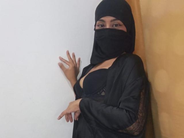LaylaNoor wearing a black hijab and mask, black satin robe, exposing her large boobs in a black lace bra, standing against a wall, hand on it.