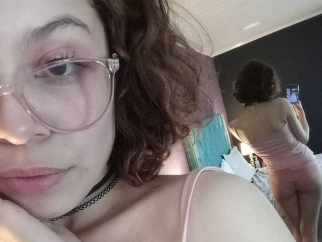Sophie Blake, con i capelli castano ricci, che indossa occhiali e un vestito rosa, mostra la schiena e i glutei in un selfie allo specchio, con una collana choker, in una camera da letto, mentre tiene in mano il telefono..