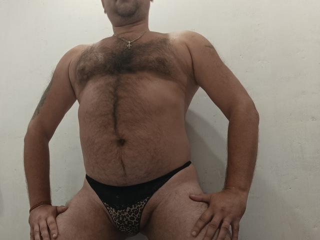 EffectGerald ist ein behaarter, muskulöser Mann mit einer Brust und einem Bauch, die mit dunklen Haaren bedeckt sind. Er trägt einen Leopardenmuster-String, sitzt mit den Händen auf den Oberschenkeln und trägt eine Halskette..