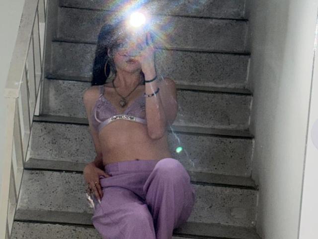 ValuuRodriguez with small tits, wearing a lavender bra and matching pants, sitting on gray stairs, flashing a camera, light flare in front, dark hair, bracelet, and necklace.
