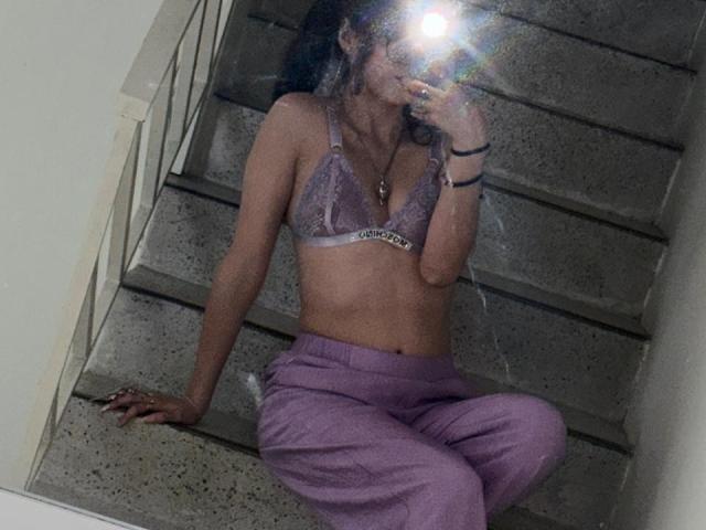 ValuuRodriguez with small tits, wearing a lilac lace bra and purple pants, sitting on concrete stairs, taking a mirror selfie with a flash obscuring her face.