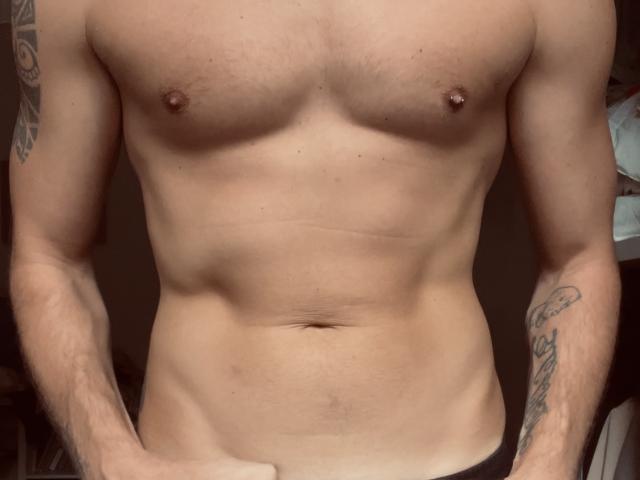 Joel69x's bare, muscular chest with small, erect nipples, tattooed arms, and a flat, toned abdomen, wearing black pants, standing with hands near waist.