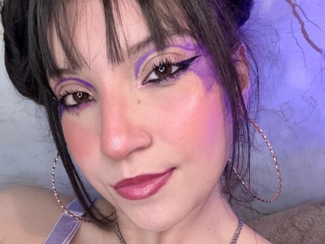 ValuuRodriguez has purple eyeshadow, black eyeliner, pinkish cheeks, and red lipstick. She's wearing hoop earrings and has dark brown hair with bangs. Her skin is fair and shiny.