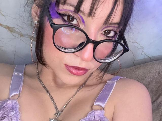 ValuuRodriguez with purple eyeshadow, black glasses, pink lipstick, purple bra, and chain necklace, looking directly at camera. Her tits are small and she has a cute face.
