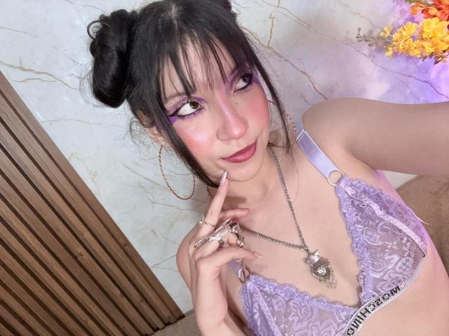 ValuuRodriguez with double buns, purple eye makeup, lavender lace bra, ring, necklace, finger on lip, winking, pink lips, pale skin, wood door, marble wall, yellow flowers.