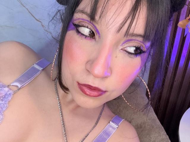 ValuuRodriguez has purple eyeshadow, pink blush, and lipstick. She's wearing a purple lace bra, large hoop earrings, and a chain necklace. Her eyes are wide, looking slightly upwards.