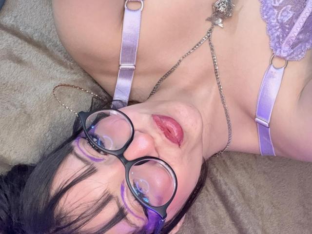 ValuuRodriguez with glasses, purple bra straps, and lipstick, wearing a chain necklace, lying on a brown surface, looking up with parted pink lips.