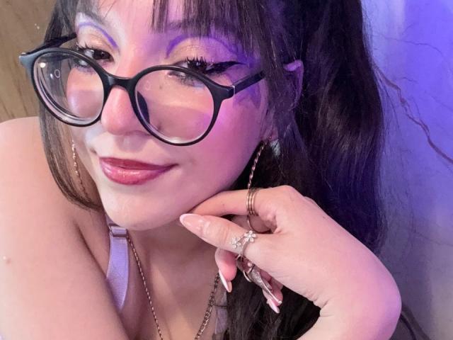 ValuuRodriguez with glasses, purple eyeshadow, and pink lipstick, smiling at the camera, showing off long nails, rings, and a necklace, with wavy black hair.
