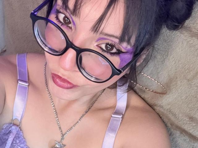ValuuRodriguez with purple eye makeup, black glasses, red lipstick, and a purple lace bra, showing off her tits, wearing a silver necklace, and a hoop earring.