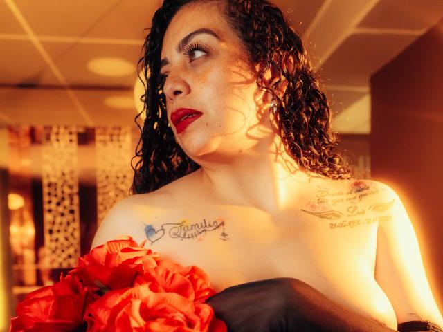 AlissaaMilf with curly black hair, red lipstick, and tattoos, holding red flowers, wearing black strapless garment, looking to the side, under warm orange light, showing her big tits.