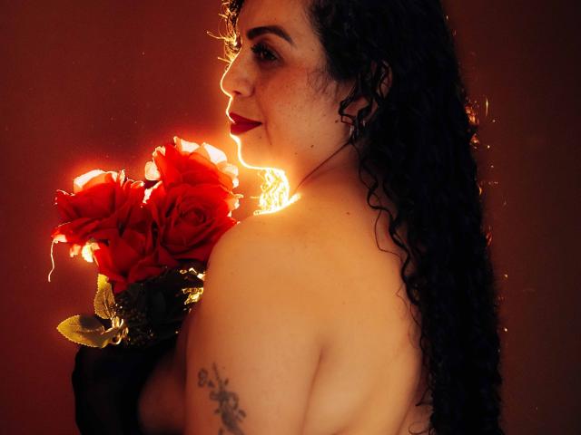 AlissaaMilf with curly black hair, red lipstick, holding red roses, back to camera, light from roses on right side, tattoo on left shoulder, soft brown background.